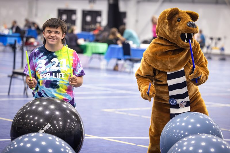 A teen boy and the Nittany Lion mascot use drumsticks to drum on exercise balls in a large gym at Penn State Health RecFest 2025. The boy is wearing a tie-dye T-shirt with the words “3.21 for Life” on it. The Nittany Lion has a scarf.