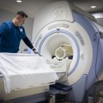 A male health care worker wearing scrubs adjusts a sheet on the table of a large MRI machine in a hospital imaging suite.