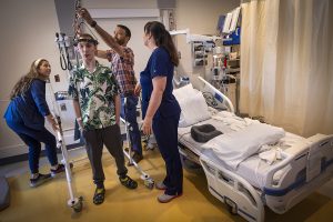 A teen boy stands wearing a halo-gravy traction device around his head. A doctor on his right adjusts the bar holding the halo up. Two nurses wearing scrubs are on the right and left of the boy and the doctor. A hospital bed and curtain are on the group’s right.