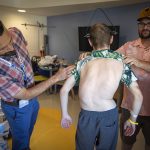 Dr. Brent O'Neill, left, leans over to look at the back of a teen boy with severe scoliosis, while the boy’s father holds the boy’s shirt up. The boy’s spine curves to the left. A halo gravity traction device is attached to his skull. They are standing in a hospital room.