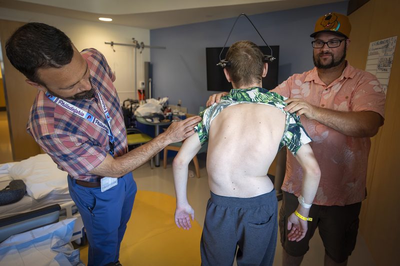 Dr. Brent O'Neill, left, leans over to look at the back of a teen boy with severe scoliosis, while the boy’s father holds the boy’s shirt up. The boy’s spine curves to the left. A halo gravity traction device is attached to his skull. They are standing in a hospital room.