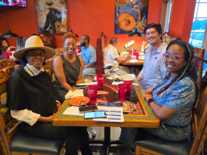 A small group of people sit at a table smiling. There are plates and cups on the table.