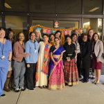 A group of diverse people standing and smiling in front of a building entryway. There is a decorative sign behind them that says, "Happy Diwali."