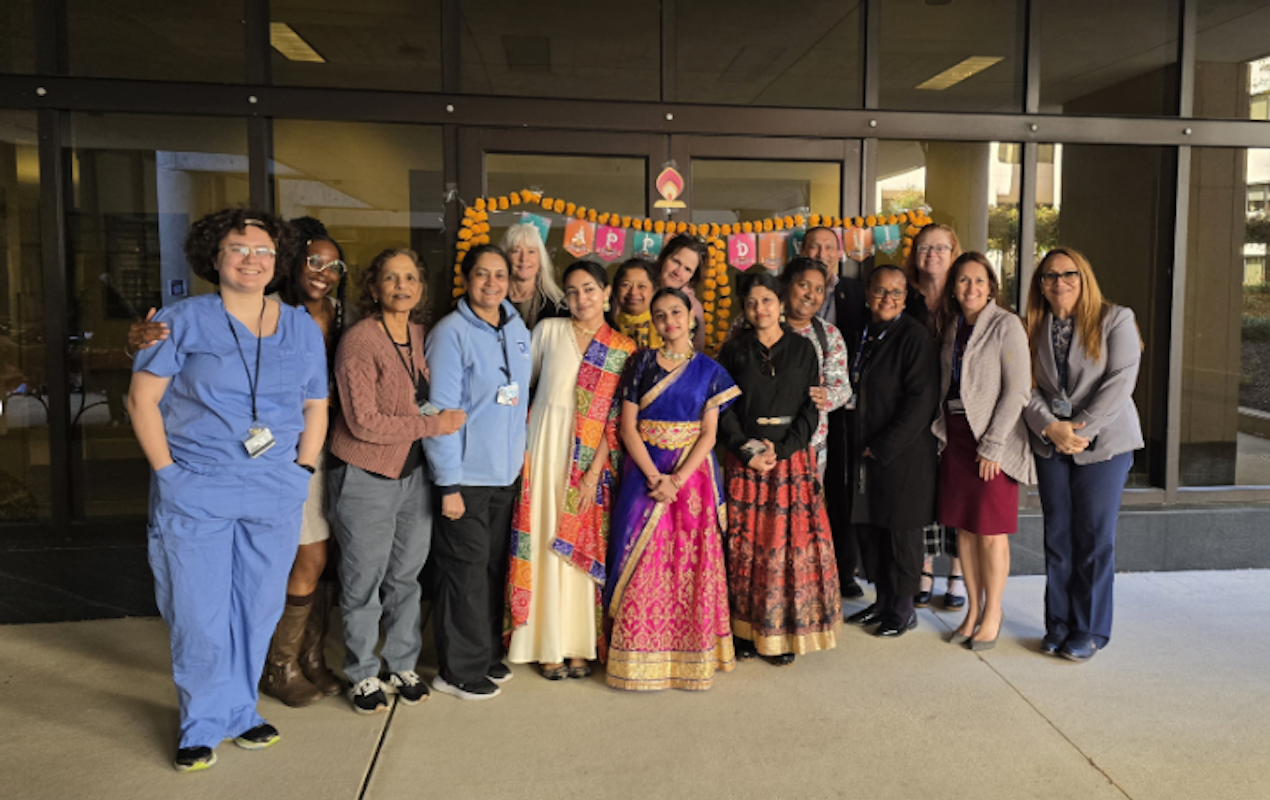 A group of diverse people standing and smiling in front of a building entryway. There is a decorative sign behind them that says,