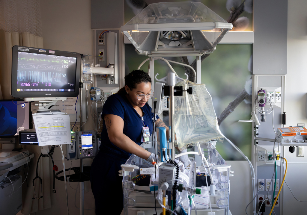 Emonie Castro, a NICU nurse, cares for a newborn in an open incubator/warmer surrounded by vital signs monitors, IV lines and bedside equipment.
