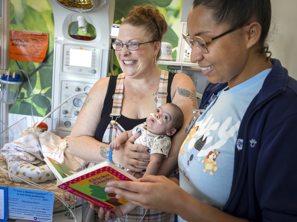 Emonie Castro, a NICU nurse wearing glasses, reads a children’s book while a smiling parent holds a baby with a small nasal tube in a NICU room.