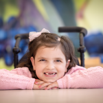 A young girl sitting at a table has her arms on the table and her chin on her arms, smiling big.