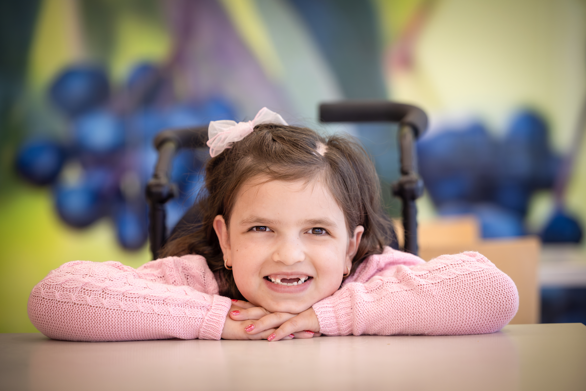 A young girl sitting at a table has her arms on the table and her chin on her arms, smiling big.