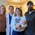 Three women and a man smile as they pose for a photo in the hallway of a doctor’s practice. From left, are Jamie Ober, registered nurse; Dr. Stephanie Estes, obstetrician-gynecologist; mother Jessica Hammaker holding a baby; and father, Justin Baer.