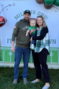 A man and woman hold their baby boy in front of a background that reads “Miles is one!” Balloons surround the background.