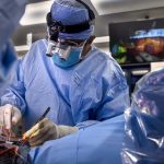 A surgeon wearing a blue surgical gown, mask, and magnifying loupes performs a heart transplant surgery in an operating room, using instruments while monitors display the procedure in the background.