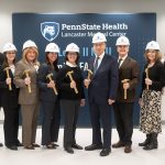 A group of hospital leaders and staff wearing white Penn State Health hard hats stand in a bright clinical room, each holding a gold ceremonial hammer. They pose smiling in front of a Penn State Health Lancaster Medical Center backdrop during a NICU groundbreaking celebration, with medical equipment visible on both sides of the room.