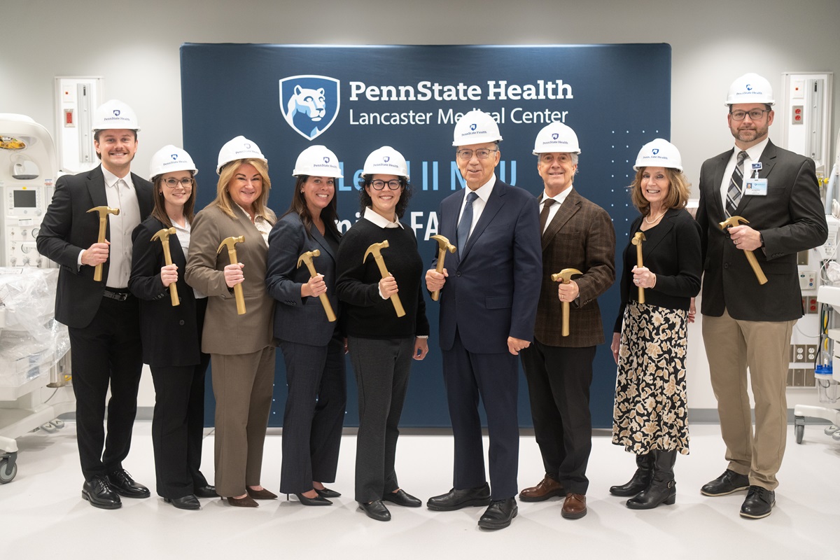 A group of hospital leaders and staff wearing white Penn State Health hard hats stand in a bright clinical room, each holding a gold ceremonial hammer. They pose smiling in front of a Penn State Health Lancaster Medical Center backdrop during a NICU groundbreaking celebration, with medical equipment visible on both sides of the room.
