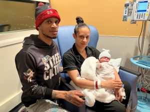 A man and woman pose with a newborn baby, who is swaddled and resting on the woman's lap. An IV pole and machine are in the background.