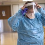A nurse dressed in a plastic gown, mask and face shield adjusts the face shield as she walks through a hospital hallway.