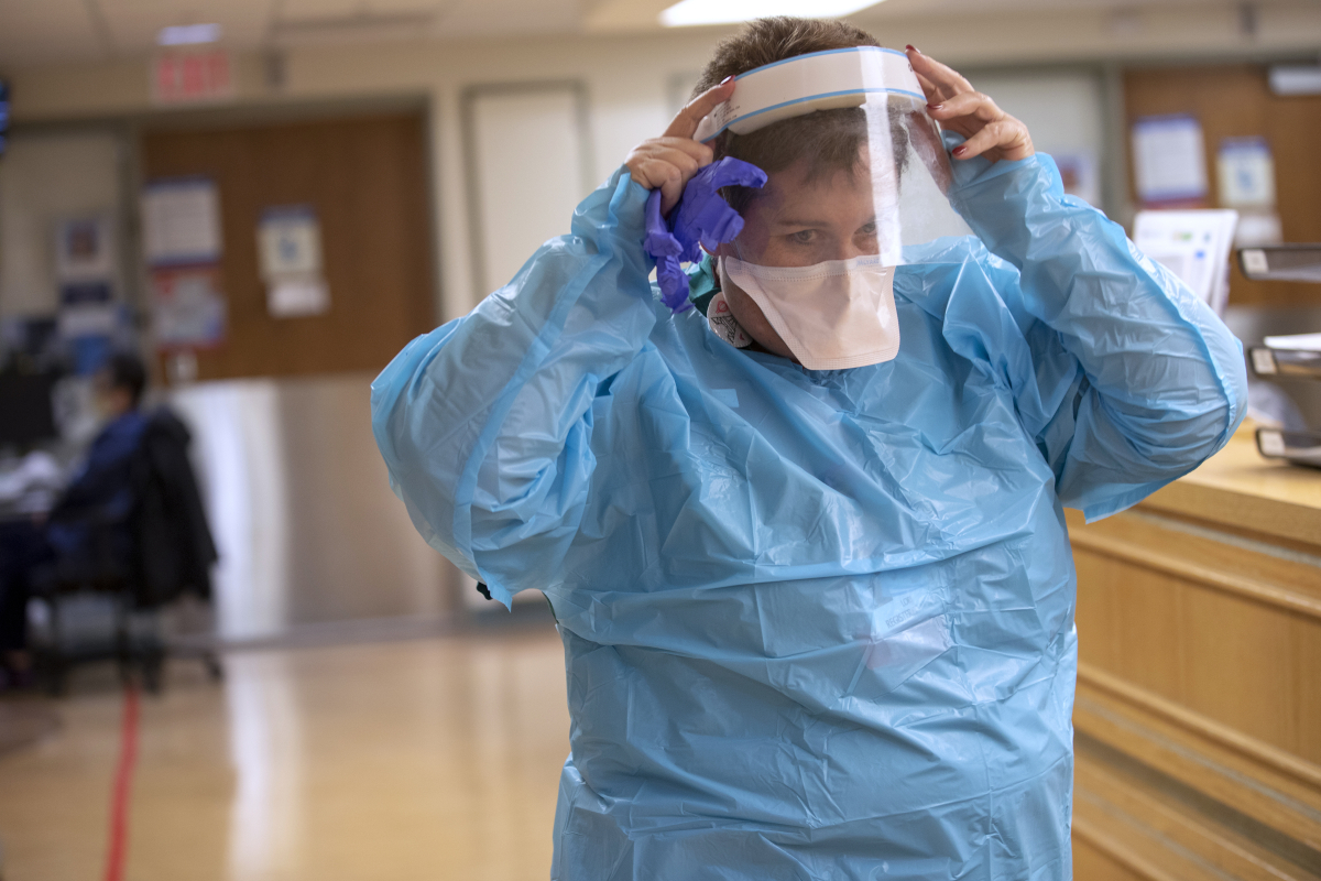 A nurse dressed in a plastic gown, mask and face shield adjusts the face shield as she walks through a hospital hallway.