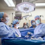 A surgical team wearing blue gowns, masks, and caps performs a procedure in a brightly lit operating room, standing around a patient draped in sterile blue sheets beneath an overhead surgical light.