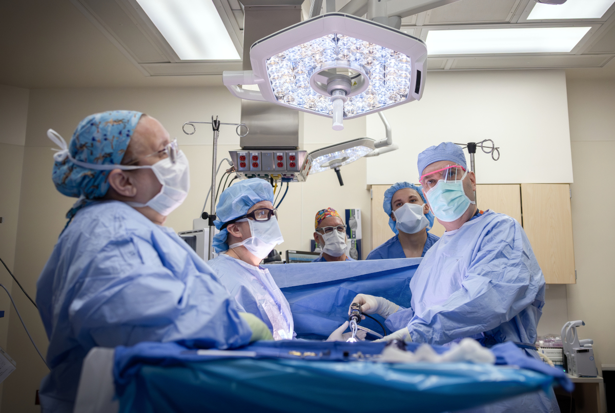A surgical team wearing blue gowns, masks, and caps performs a procedure in a brightly lit operating room, standing around a patient draped in sterile blue sheets beneath an overhead surgical light.