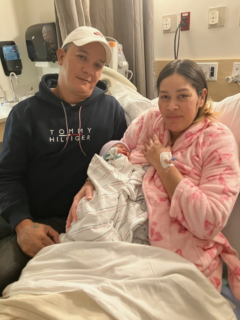 Two people pose for a photo, as one of them - a woman in a hospital bed - holds a newborn child.
