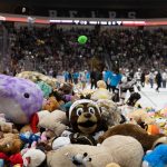 Hundreds of stuffed animals cover the ice during a hockey arena’s teddy bear toss, with players and staff on the rink and a packed crowd watching in the stands.