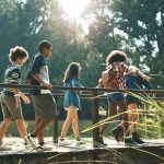 Five children walk across a wooden walking bridge. Various trees and weeds are in the foreground and background.