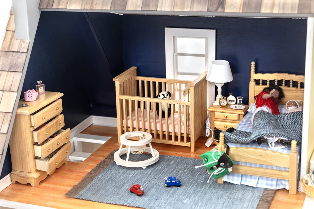 A dollhouse bedroom shows a mother sleeping with a baby, a walker on the floor and a stuffed animal in a crib.