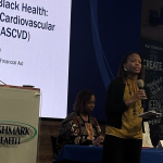 A woman stands on a stage speaking into a microphone. There is a table behind her with people sitting at it. There are projection screens behind her and a podium to the side.