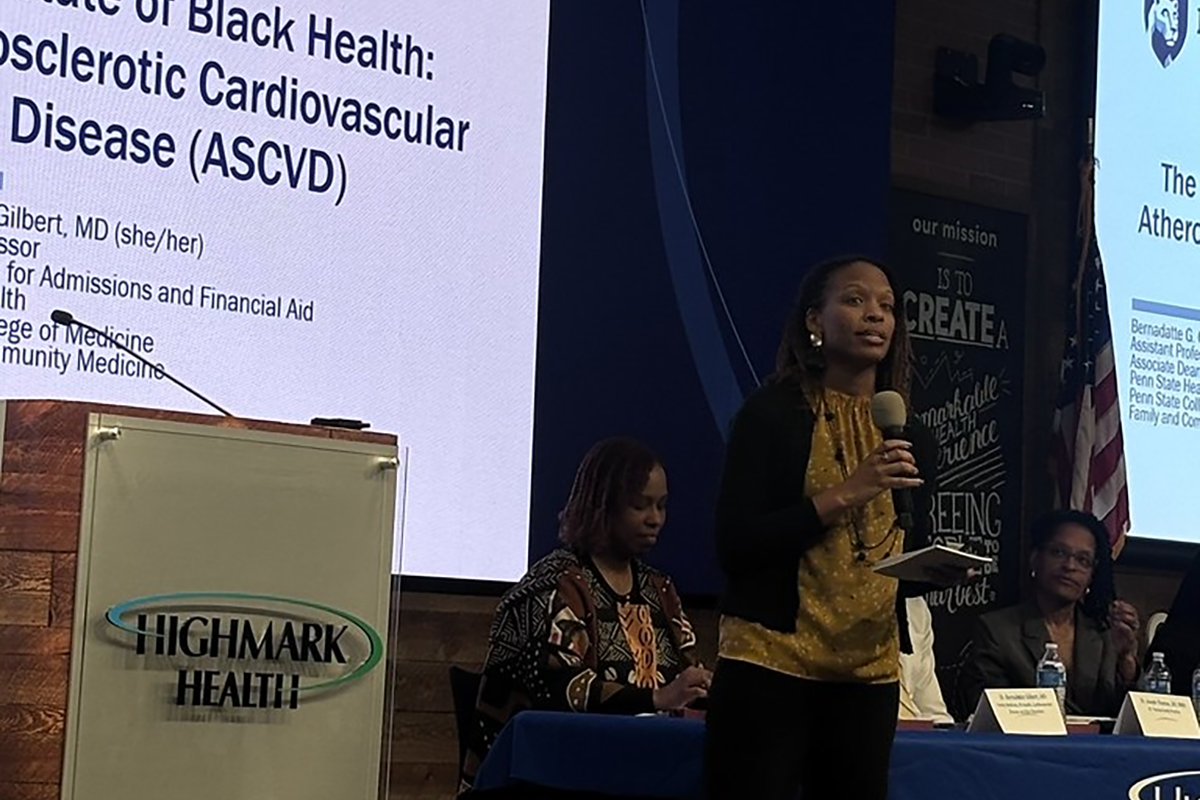 A woman stands on a stage speaking into a microphone. There is a table behind her with people sitting at it. There are projection screens behind her and a podium to the side.
