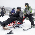 A woman is seated on an adaptive sled, while a ski instructor in winter clothing and on skis holds onto the back of the sled. They are on snow, and a few people look on from the background.