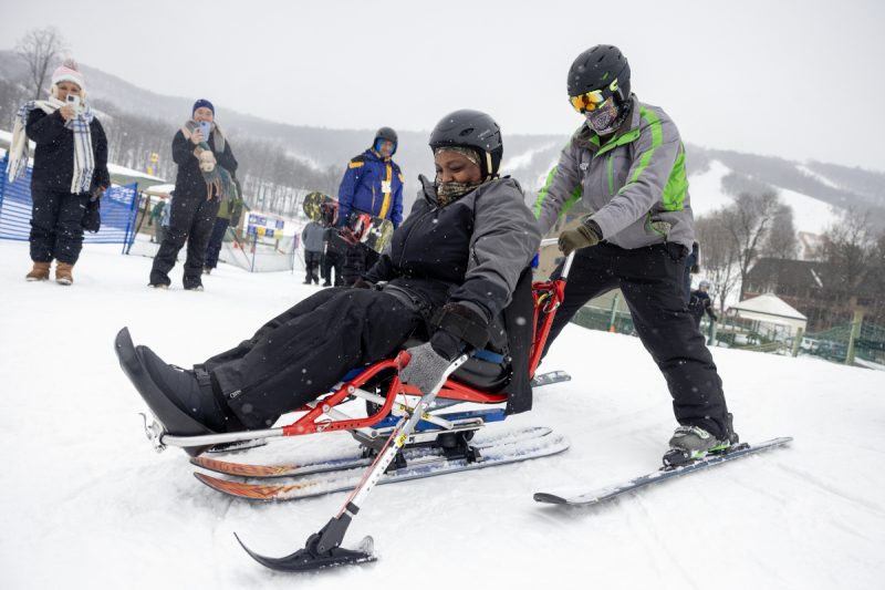 A woman is seated on an adaptive sled, while a ski instructor in winter clothing and on skis holds onto the back of the sled. They are on snow, and a few people look on from the background.