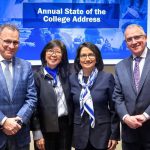 Two men and two women wearing business attire stand side-by-side, smiling. Behind them is a sign that says, "Annual State of the College Address."