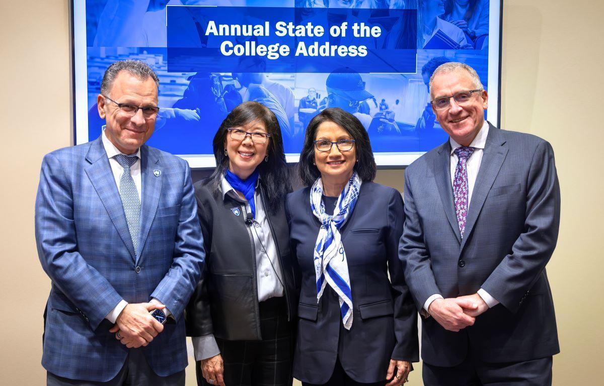 Two men and two women wearing business attire stand side-by-side, smiling. Behind them is a sign that says, 