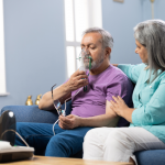 An older gentleman sits on a couch and holds an oxygen mask over his mouth and nose, holding his eyeglasses with his other hand. A woman of similar age sits beside him and puts her arm around his shoulders.