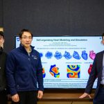 Three men pose for a photo in front of a television that reads "Self-organizing Heart Modeling and Simulation" with a series of graphics showing the heart in different phases of self-organization, with electrical maps of a heart beneath it.