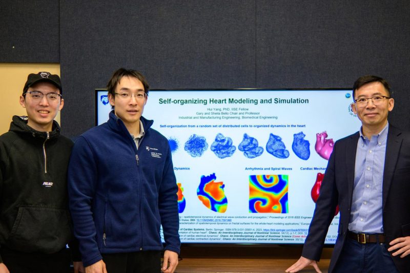 Three men pose for a photo in front of a television that reads "Self-organizing Heart Modeling and Simulation" with a series of graphics showing the heart in different phases of self-organization, with electrical maps of a heart beneath it.