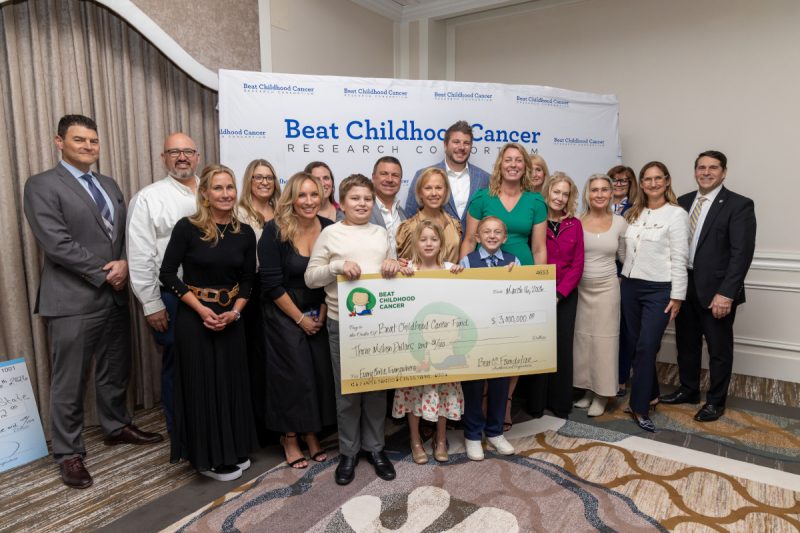 A group of adults and children pose with an oversized check in the amount of $3 million. A backdrop contains the Beat Childhood Cancer Research Consortium logo.
