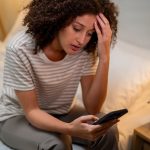 Stressed woman uses mobile phone as she sits on bed at night.