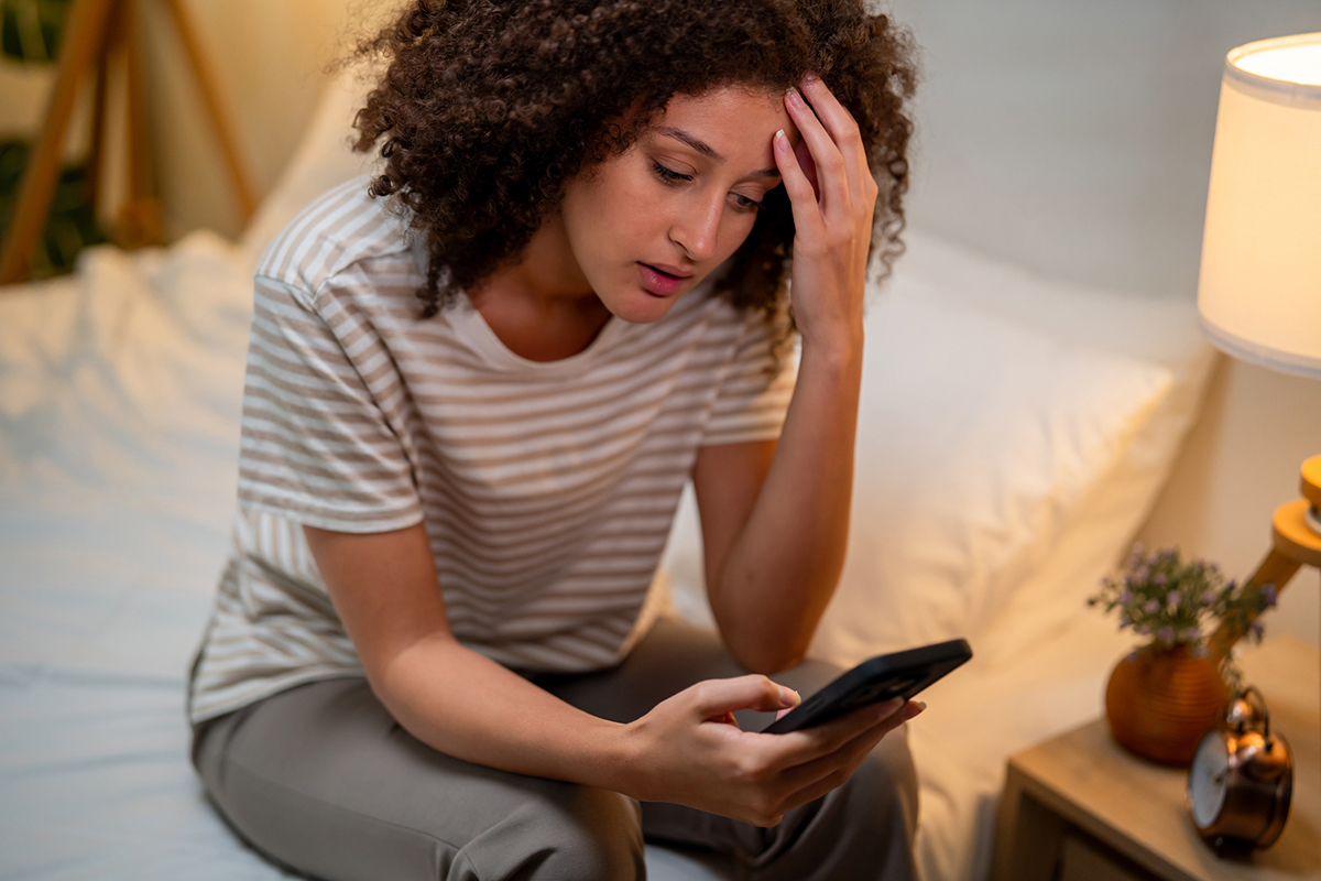 Stressed woman uses mobile phone as she sits on bed at night.