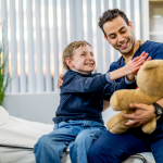 A male clinician wearing scrubs sits next to a young boy who seems to be handicapped. The gentleman is holding a teddy bear and the boy is reaching for it. They are both smiling.