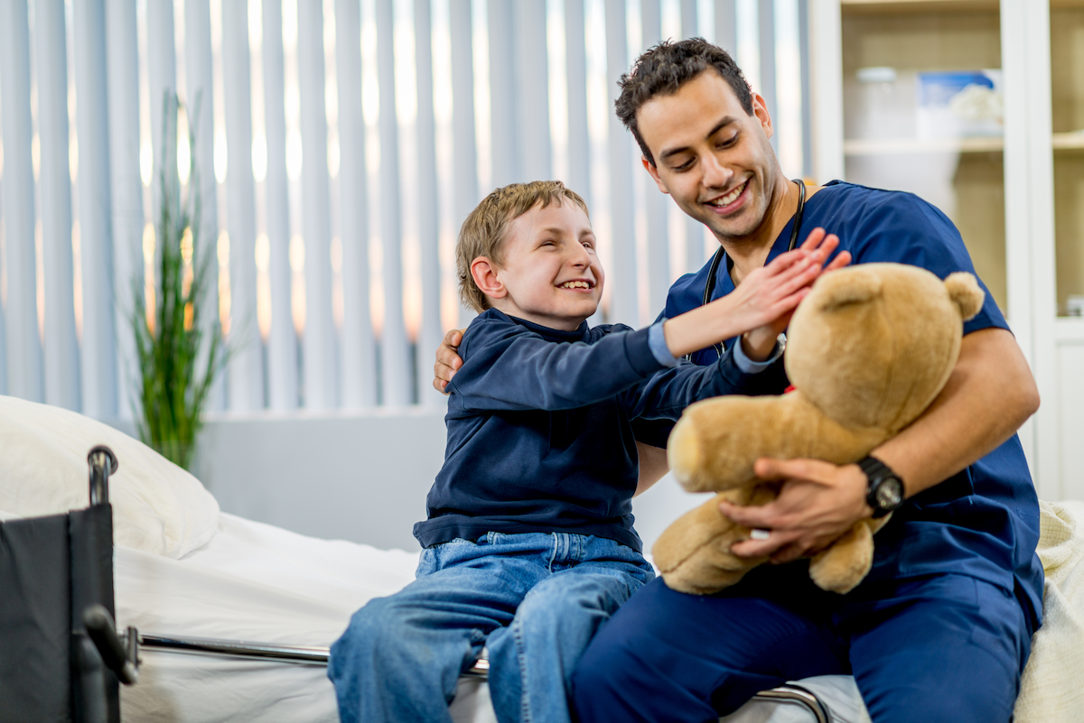 A male clinician wearing scrubs sits next to a young boy who seems to be handicapped. The gentleman is holding a teddy bear and the boy is reaching for it. They are both smiling.