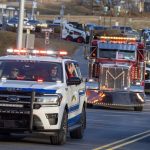 A line of vehicles with flashing lights drives toward the camera. They include emergency vehicles, tow trucks and others.