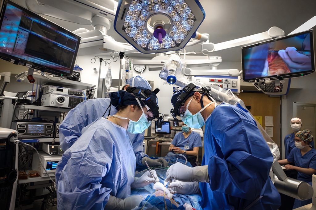 A surgeon, surgical resident and surgical assistant wearing surgical gowns and masks perform surgery on a patient at Penn State Health Milton S. Hershey Medical Center. Two monitors are on the left and right side of the room, and a light is above them. 