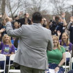 A gentleman is a business suit facing away addresses an audience seated in lawn chairs and standing. The people in the audience have their hands raised.