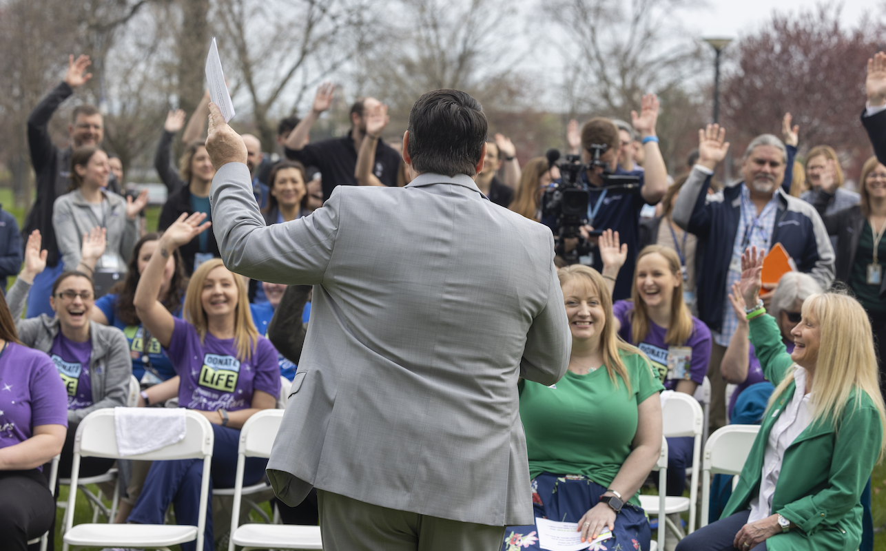 A gentleman is a business suit facing away addresses an audience seated in lawn chairs and standing. The people in the audience have their hands raised.