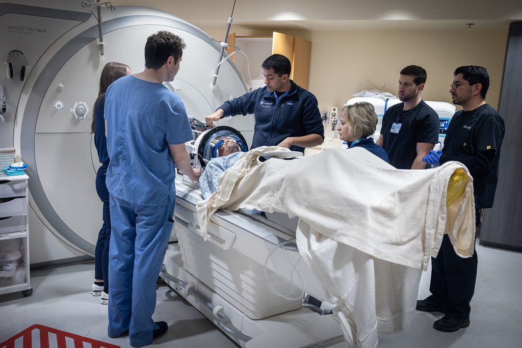 A group of five MRI technologists and one physician stand next to a male patient who is about to go into an MRI machine. The physician, center, points to the patient.