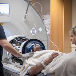 Two MRI technologists stand next to a male patient who is about to go into an MRI machine. The patient has a headgear on his head and a nasal cannula under his nose.