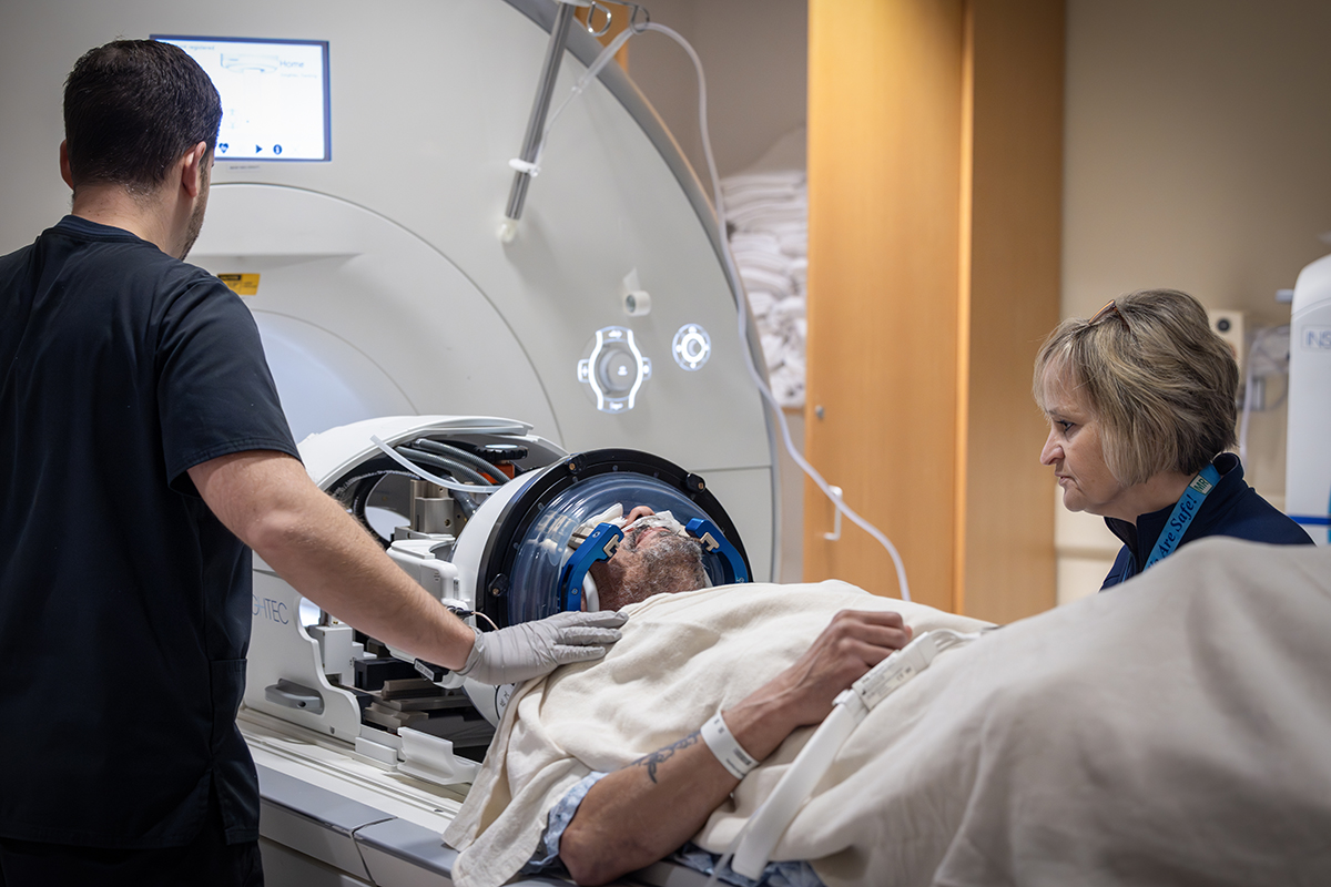 Two MRI technologists stand next to a male patient who is about to go into an MRI machine. The patient has a headgear on his head and a nasal cannula under his nose.