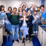 Ten people in a lab pose for a photo shaping their fingers into a triangle