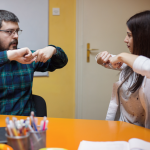A young man wearing glasses and a plaid shirt sits on the left of the image using his hands to sign a word in American Sign Language, while an ASL teacher, a young woman in a blouse and sweater who is sitting on the right, confirms his sign by demonstrating the same hand gesture.