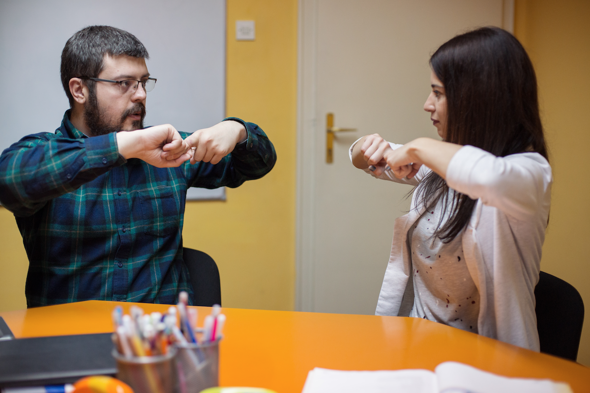 A young man wearing glasses and a plaid shirt sits on the left of the image using his hands to sign a word in American Sign Language, while an ASL teacher, a young woman in a blouse and sweater who is sitting on the right, confirms his sign by demonstrating the same hand gesture.